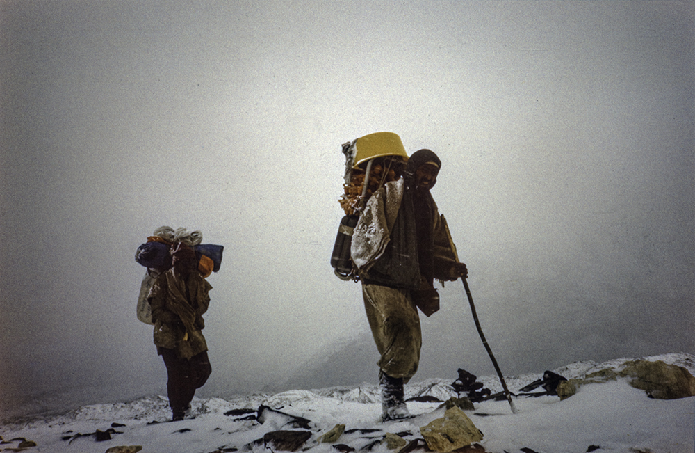 In the foreground a porter carrying a package of chicken eggs on his back.